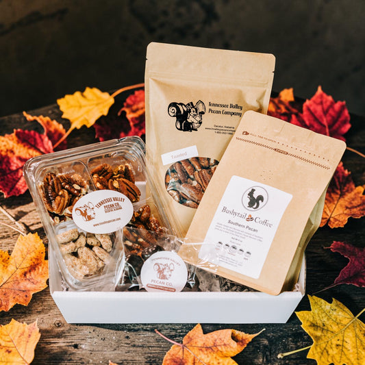 A fall-themed gift basket containing a bag of Tennessee Valley Pecan Company pecans, a bag of Bushy Tail Coffee medium roast, and a clear container of mixed pecans, arranged in a white basket with autumn leaves on a rustic wooden surface.