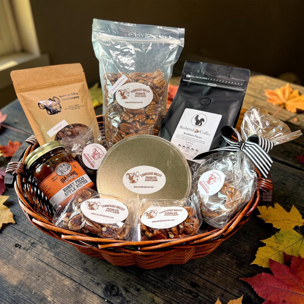 A fall-themed gourmet gift basket filled with various packaged snacks and treats, including pecans, coffee, and pecan butter, arranged on a rustic wooden table with scattered autumn leaves.