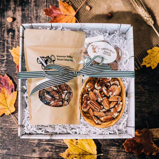 A gift box of fall treats from Tennessee Valley Pecan Company, featuring a kraft paper bag of pecans tied with a striped ribbon, a small bowl of pecans, and a packaged chocolate, arranged in white paper shred on a rustic wooden background with scattered autumn leaves.