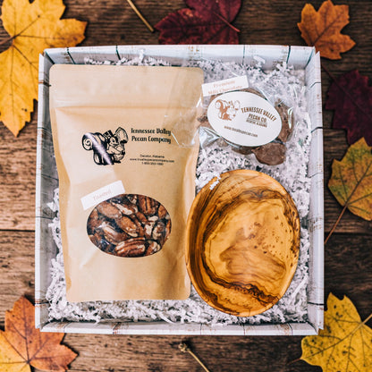 A gift box of Tennessee Valley Pecan Company products, including a bag of pecans, a small bag of milk chocolate pecans, and a wooden serving bowl, arranged in white paper shred on a rustic wooden background with autumn leaves.