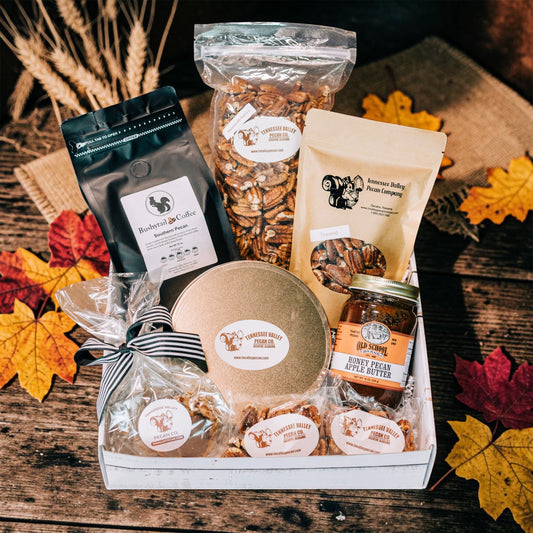 A fall-themed gourmet gift basket filled with various packaged snacks and treats, including roasted coffee beans, honey, and dried pecans, arranged in a white wooden box on a rustic wooden table with autumn leaves.
