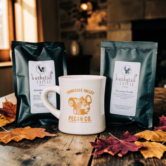 Two bags of Bushy Tail Coffee and a white ceramic mug printed with Tennessee Valley Pecan Co. branding, arranged on a rustic wooden table with autumn leaves in a warm indoor setting.