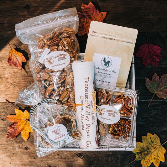 A gift basket filled with packaged food items, including a large bag of pecans, a bag of Tennessee Valley Pecan Co. pecans, a bag of Babytail Coffee roasted flaxseed flour, and a tea towle, arranged on a rustic wooden surface with scattered autumn leaves.