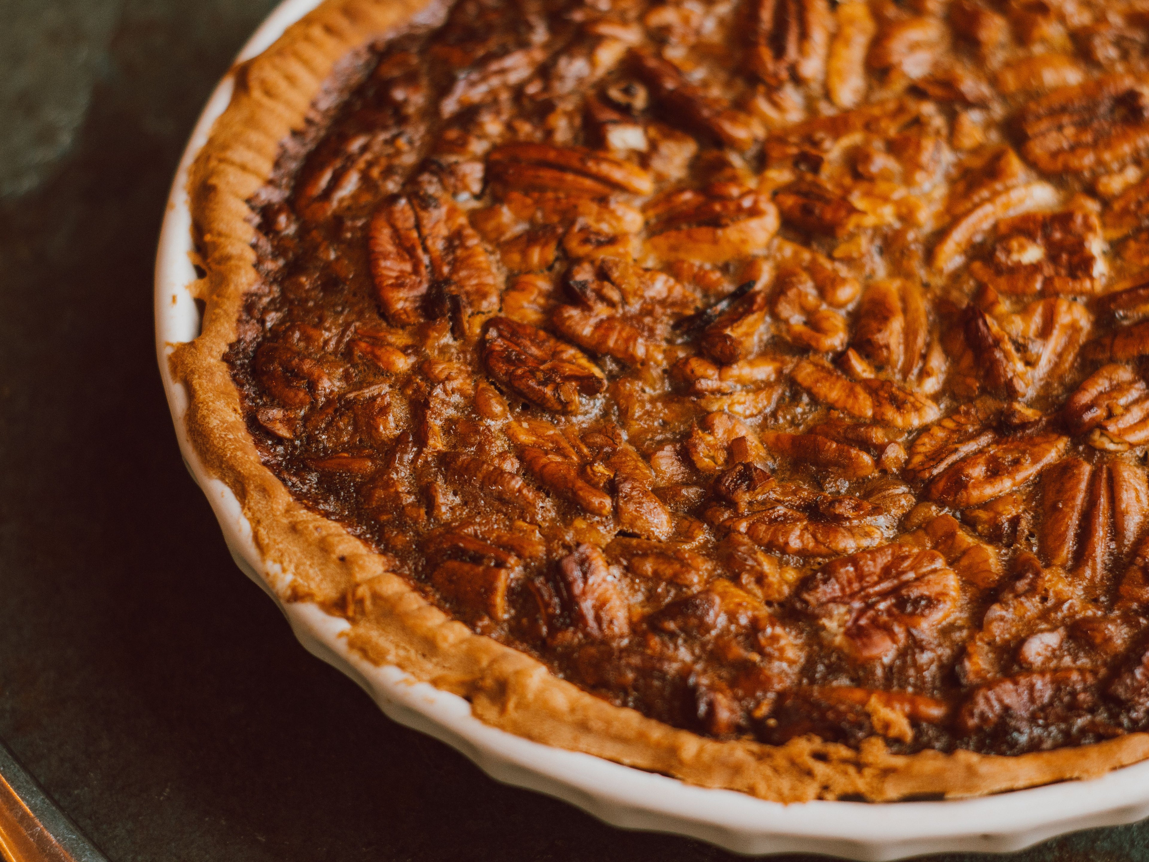A close-up shot of a freshly baked pecan pie in a white ceramic dish, showing the golden-brown crust and glossy pecan topping.
